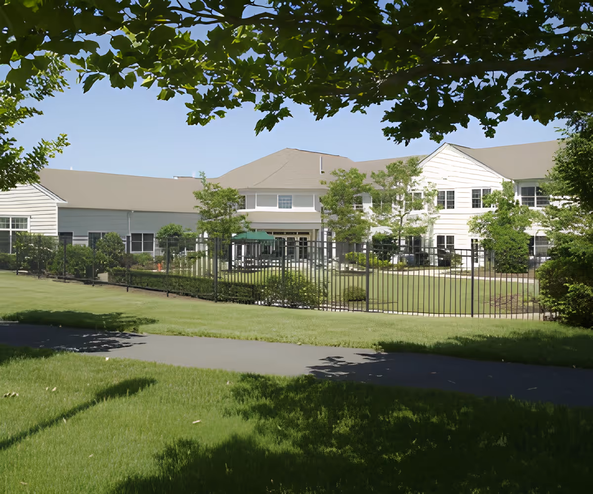 Front exterior of a two-story senior living facility with a fenced lawn, trees, and a sidewalk in the foreground.