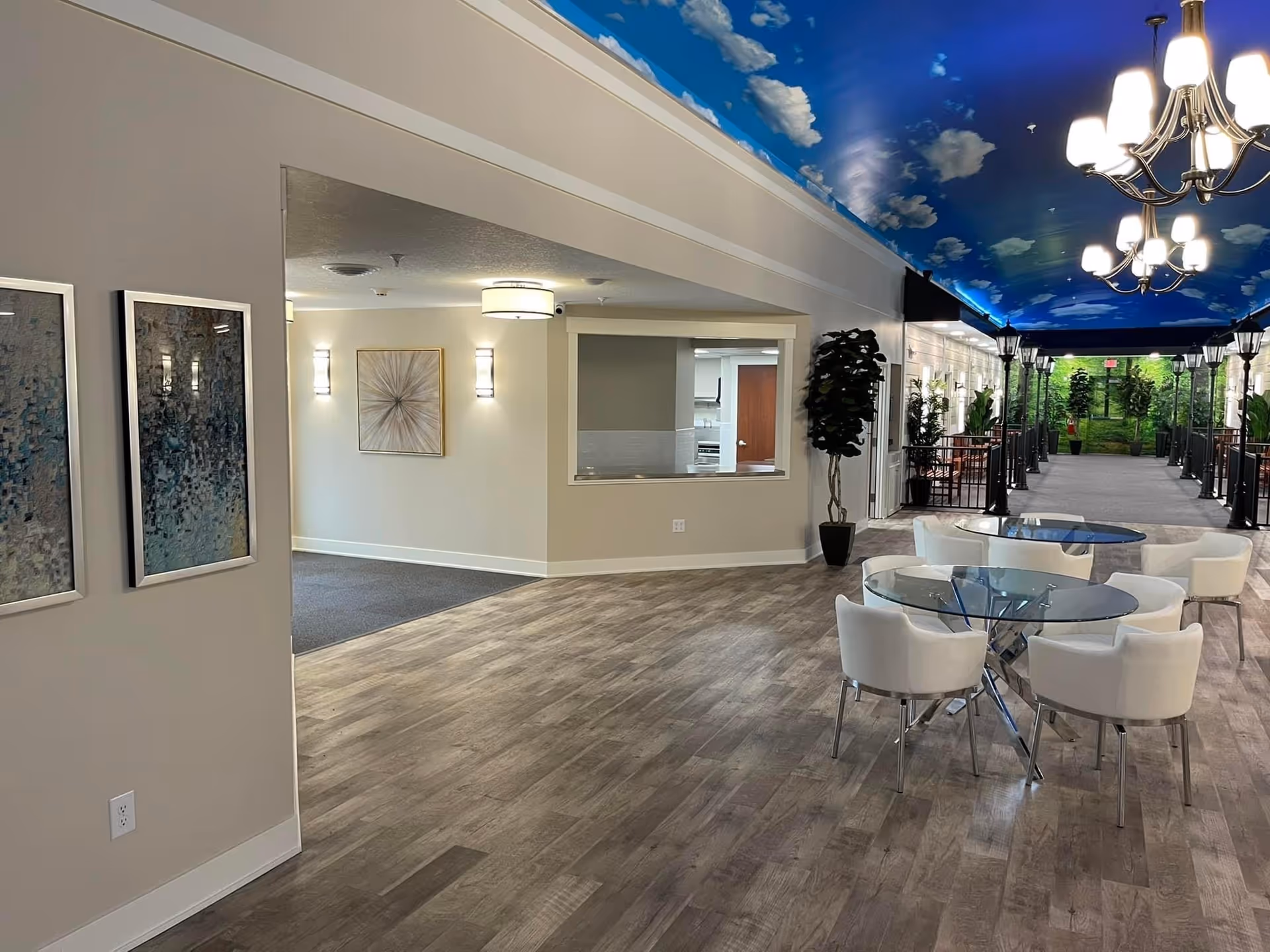 Interior view of a senior living facility with a hallway featuring a ceiling painted like a blue sky with clouds. There are glass tables with white chairs on the right side, potted plants along the hallway, and wall art on the left. The floor is wood-patterned, and the area is well-lit with modern light fixtures.