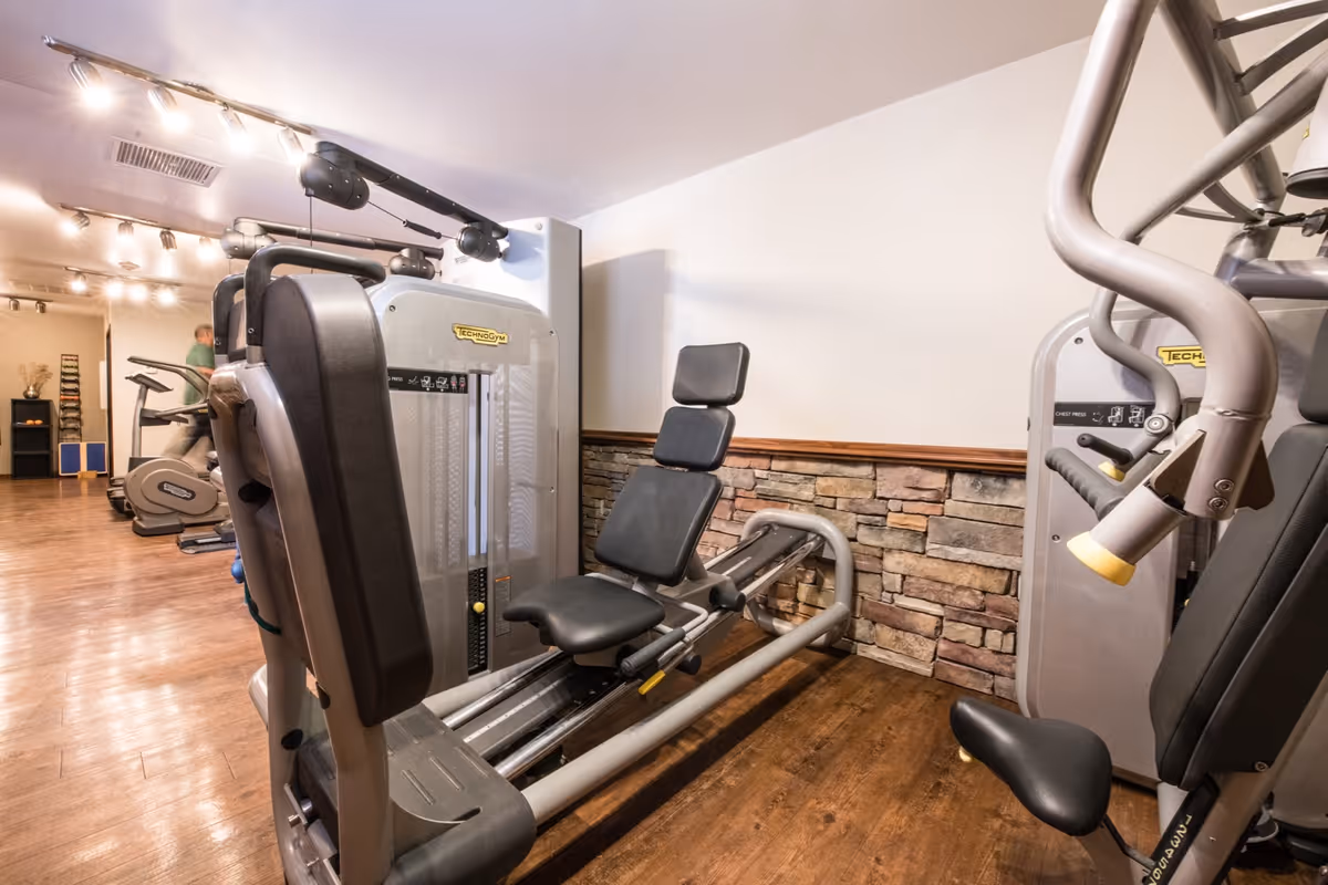 Interior fitness room with strength-training machines and cardio equipment against a stone-accent wall and wood flooring.