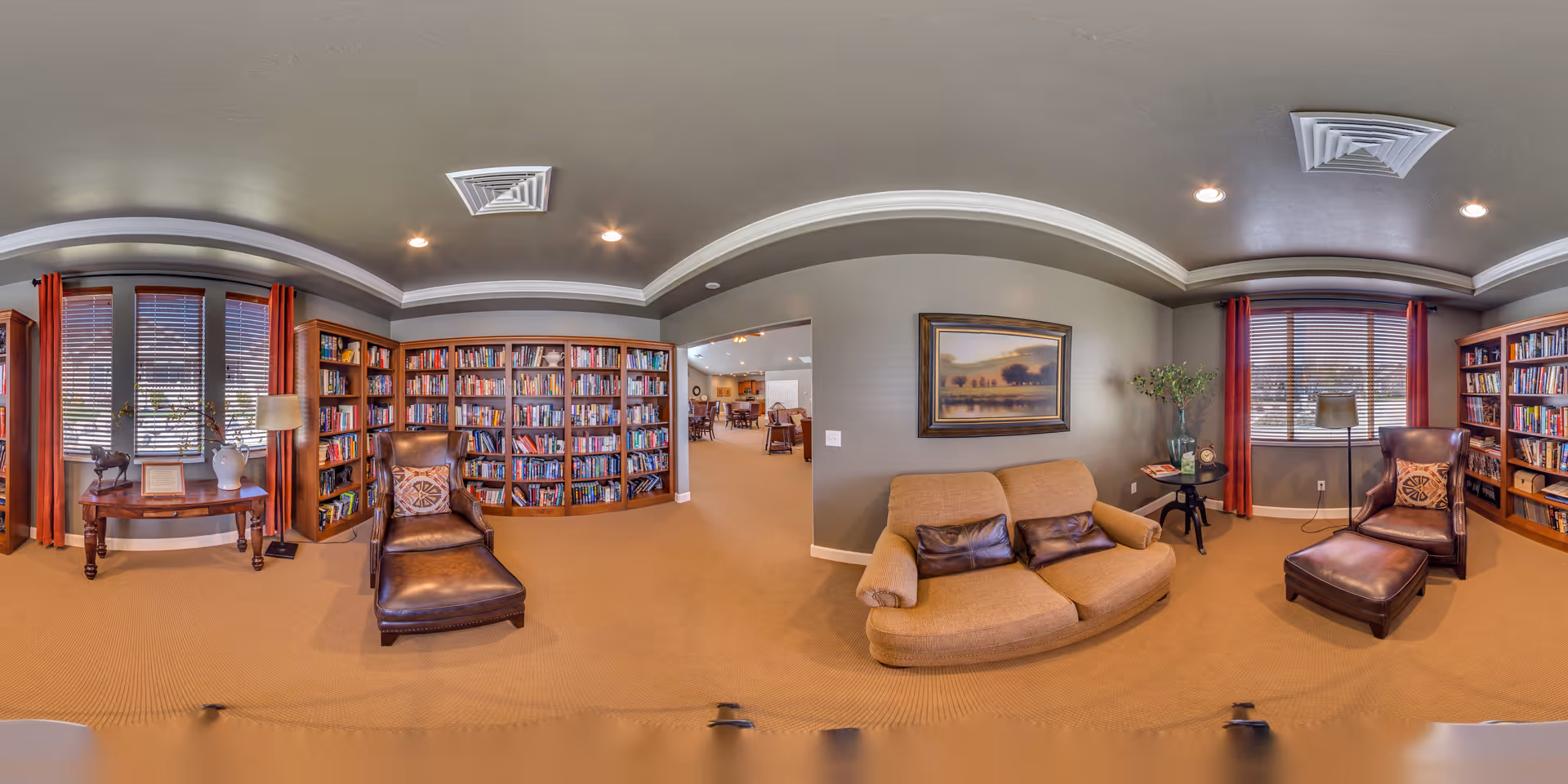 A cozy library room with large bookshelves filled with books along the walls, two brown leather armchairs with matching ottomans, a beige loveseat with two brown pillows, a wooden side table with a lamp and decorative items, and windows with red curtains allowing natural light to enter.