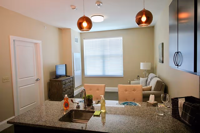 Interior view of a senior living facility apartment showing a kitchen island with a sink, soap dispensers, and a small plant. Behind the island are two cushioned chairs, a living area with a sofa, a side table with a lamp, a TV on a wooden dresser, and a window with blinds. The walls are painted beige, and there are two pendant lights hanging from the ceiling.