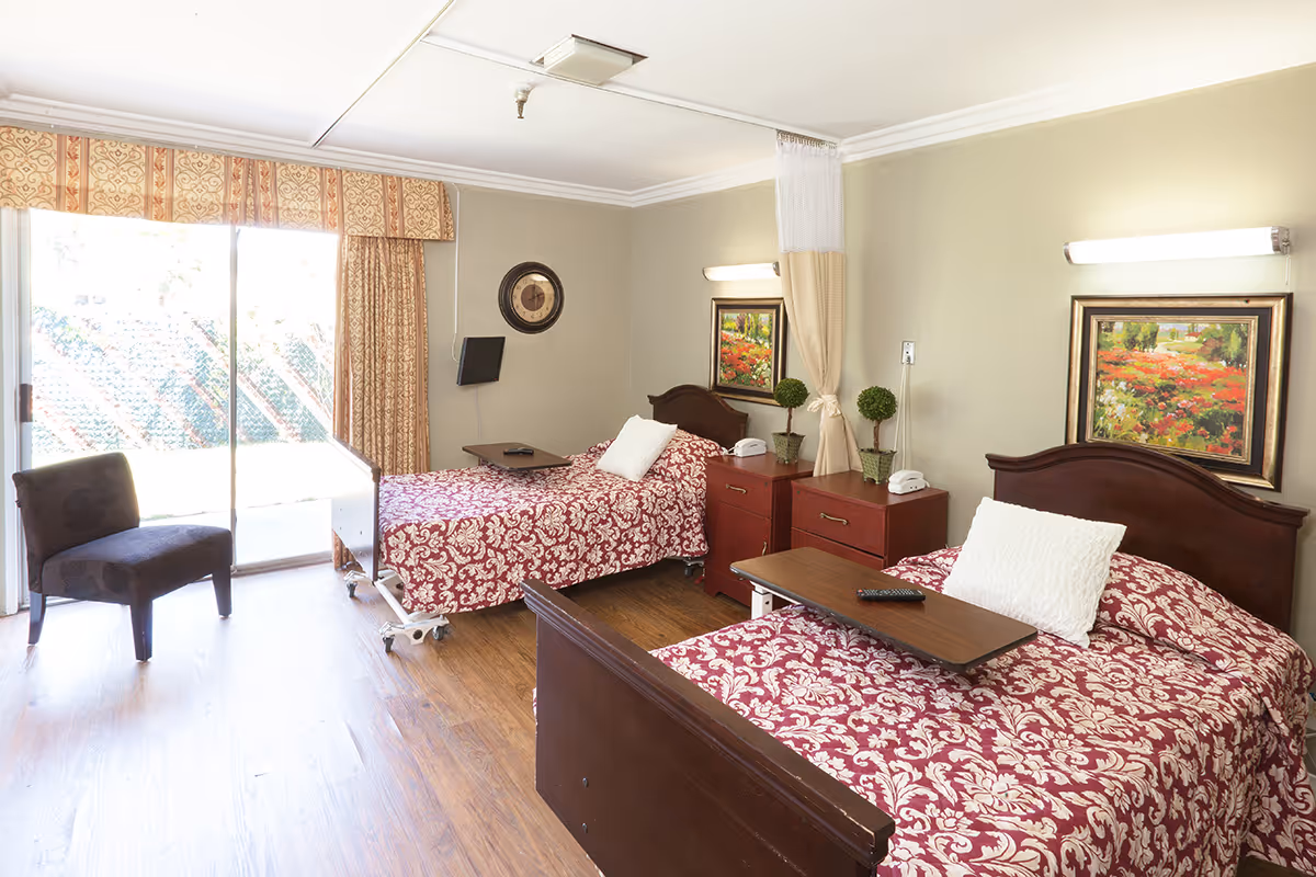 A bright and clean bedroom in a healthcare facility featuring two single beds with red and white patterned bedspreads, each with a white pillow and a wooden overbed table. The room has wooden flooring, a dark upholstered chair near a sliding glass door with patterned curtains, two bedside tables with telephones and small potted plants, a wall clock, and framed floral paintings on the walls.
