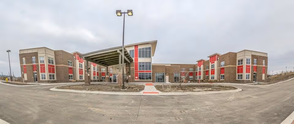Wide exterior view of The Healthcare Resort of Olathe building with a modern design featuring large windows, red and beige panels, and a covered entrance area. The surrounding area includes paved roads and minimal landscaping under an overcast sky.