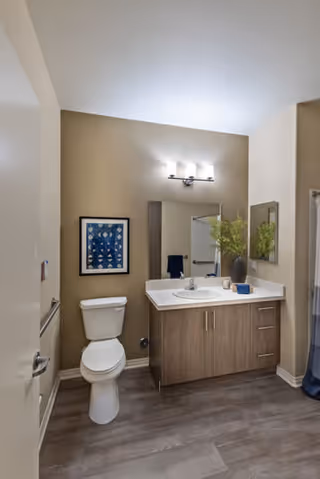 A bathroom with a white toilet and a wooden vanity with a white countertop. Above the vanity is a mirror and a light fixture with three bulbs. There is a framed artwork on the wall next to the toilet and a vase with green plants on the countertop. The floor has wood-like tiles and the walls are painted beige.