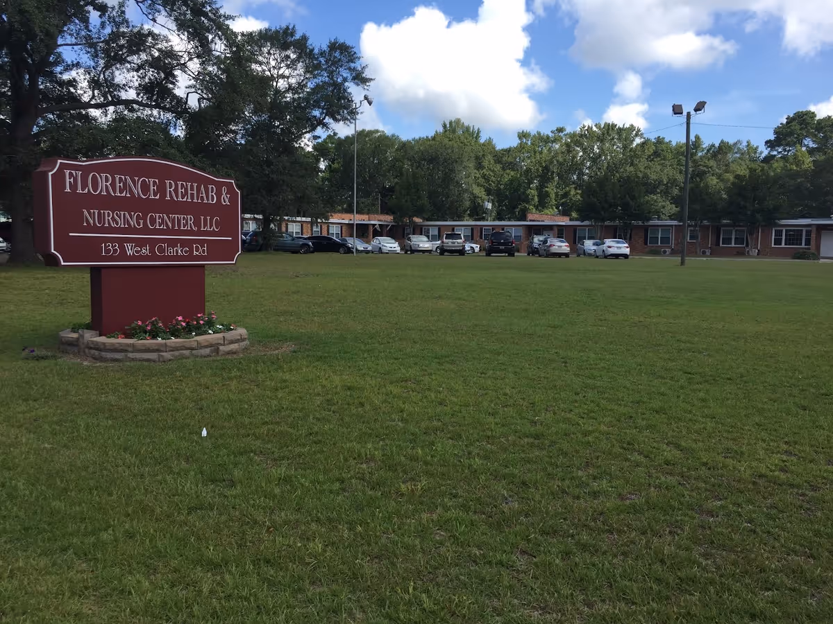 A large grassy lawn with a maroon sign that reads 'FLORENCE REHAB & NURSING CENTER, LLC 133 West Clarke Rd' in front of a single-story brick building with multiple windows and parked cars. Trees and a partly cloudy sky are visible in the background.