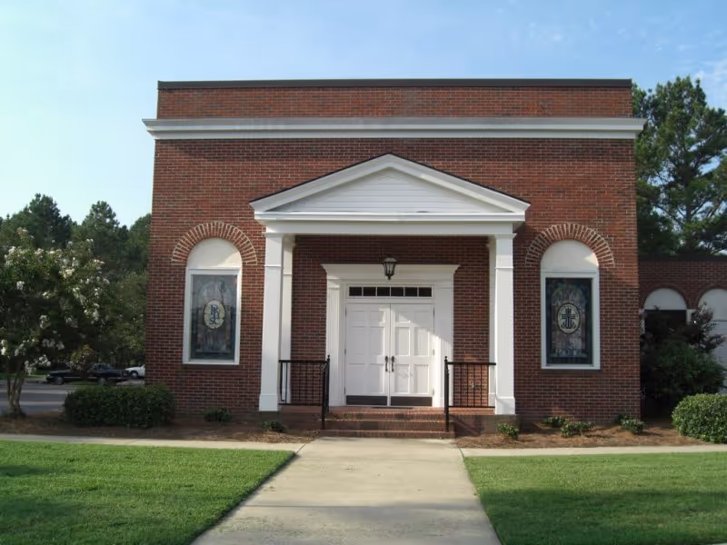 Brick building facade with a white double-door entrance under a pedimented portico, flanked by stained-glass windows and a front walkway and lawn.