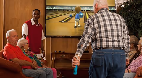 A group of elderly people seated in a living room area watching a man play a virtual bowling game on a large screen. A staff member stands nearby observing. The room has wood-paneled walls and comfortable seating.
