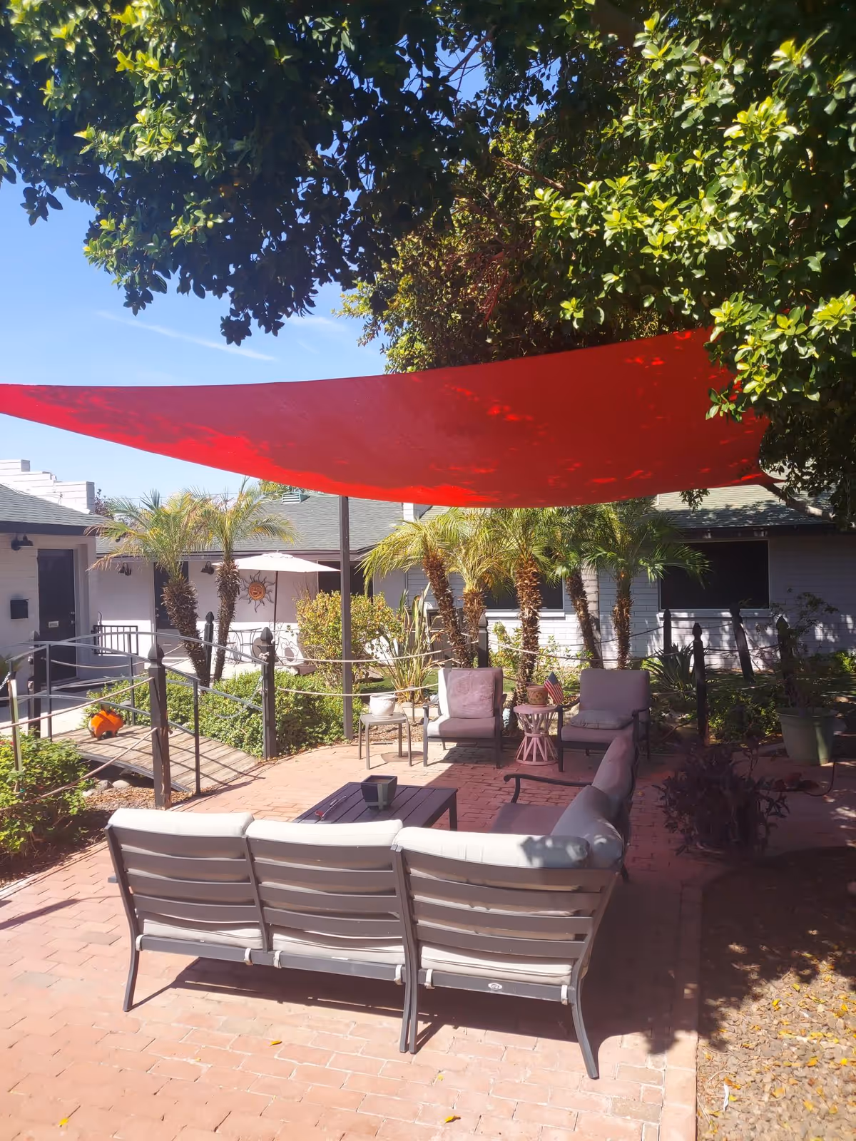 Outdoor courtyard with patio seating under a red shade sail, surrounded by palm plants and building walkways.
