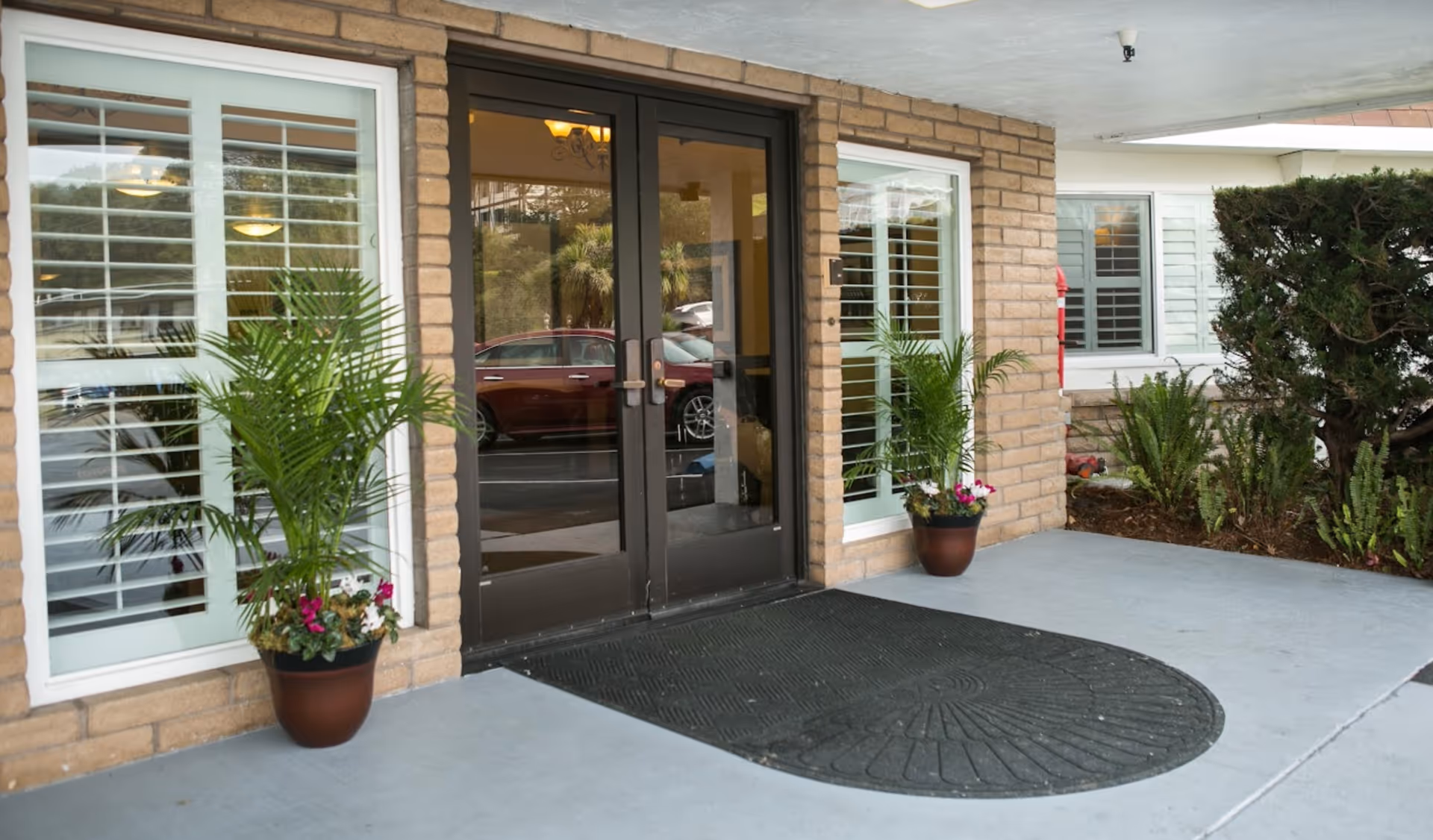 Front entrance of a brick building with double glass doors, potted plants, shuttered windows, and a large entrance mat.