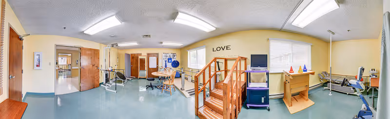 Rehabilitation therapy room showing parallel bars, exercise equipment, a wooden practice staircase, table and chairs under fluorescent lighting.