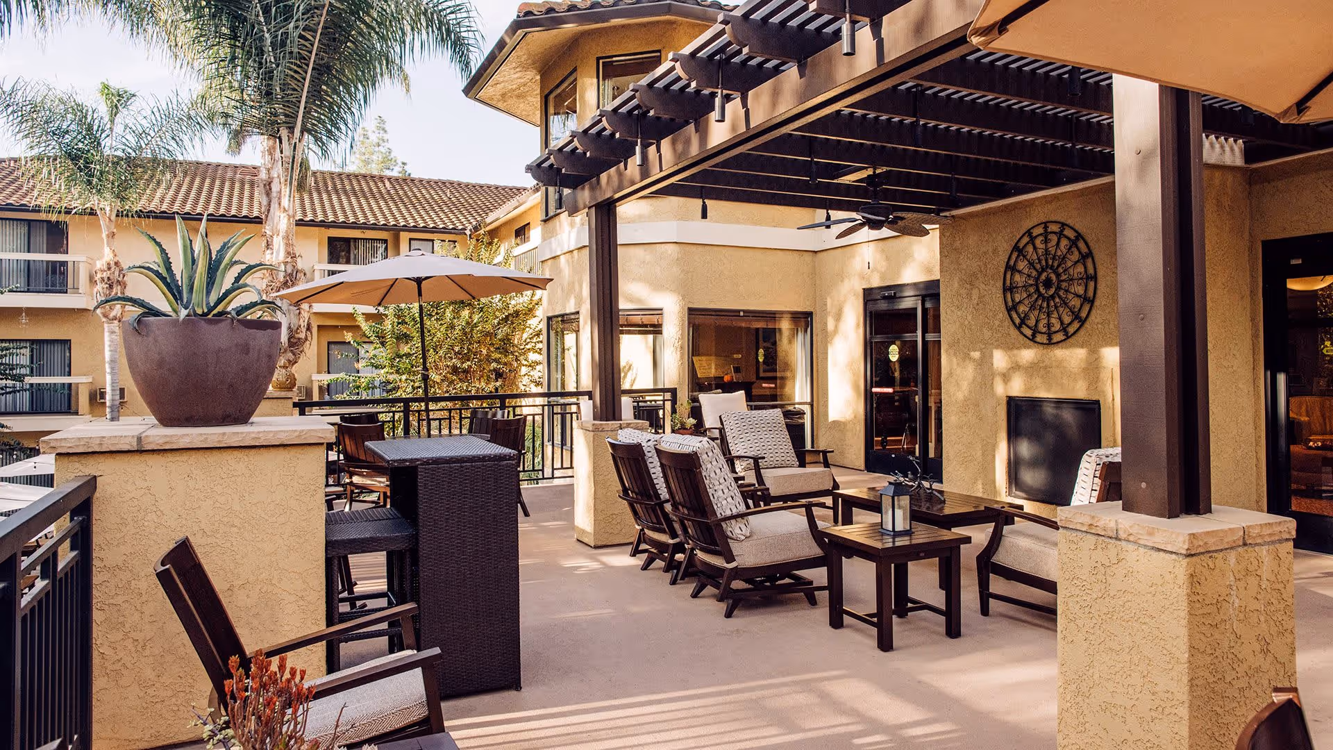 Outdoor patio area at a senior living facility with cushioned chairs, tables, a large potted plant, an umbrella, and a pergola overhead. The building exterior is visible with windows and balconies, surrounded by palm trees.