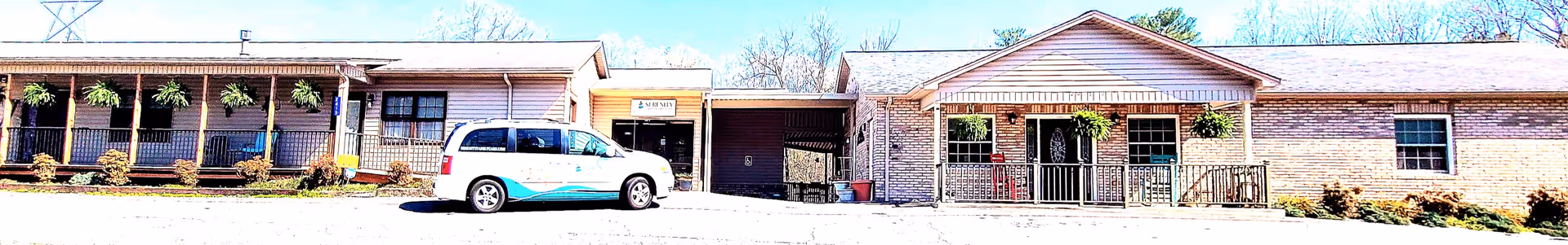 Exterior front view of a single-story brick building with a covered porch, several windows, and a ramp leading to the entrance. A white van with blue graphics is parked in front of the building under a clear sky.