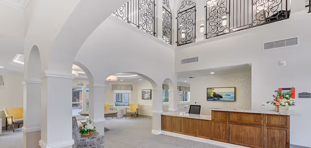 A bright and spacious senior living facility lobby with white walls, arched doorways, and a wooden reception desk. The area features comfortable seating with yellow and gray chairs, decorative wrought iron railings on the upper level, and a large landscape painting on the wall behind the desk. There are flowers on the reception desk and natural light coming through the windows.