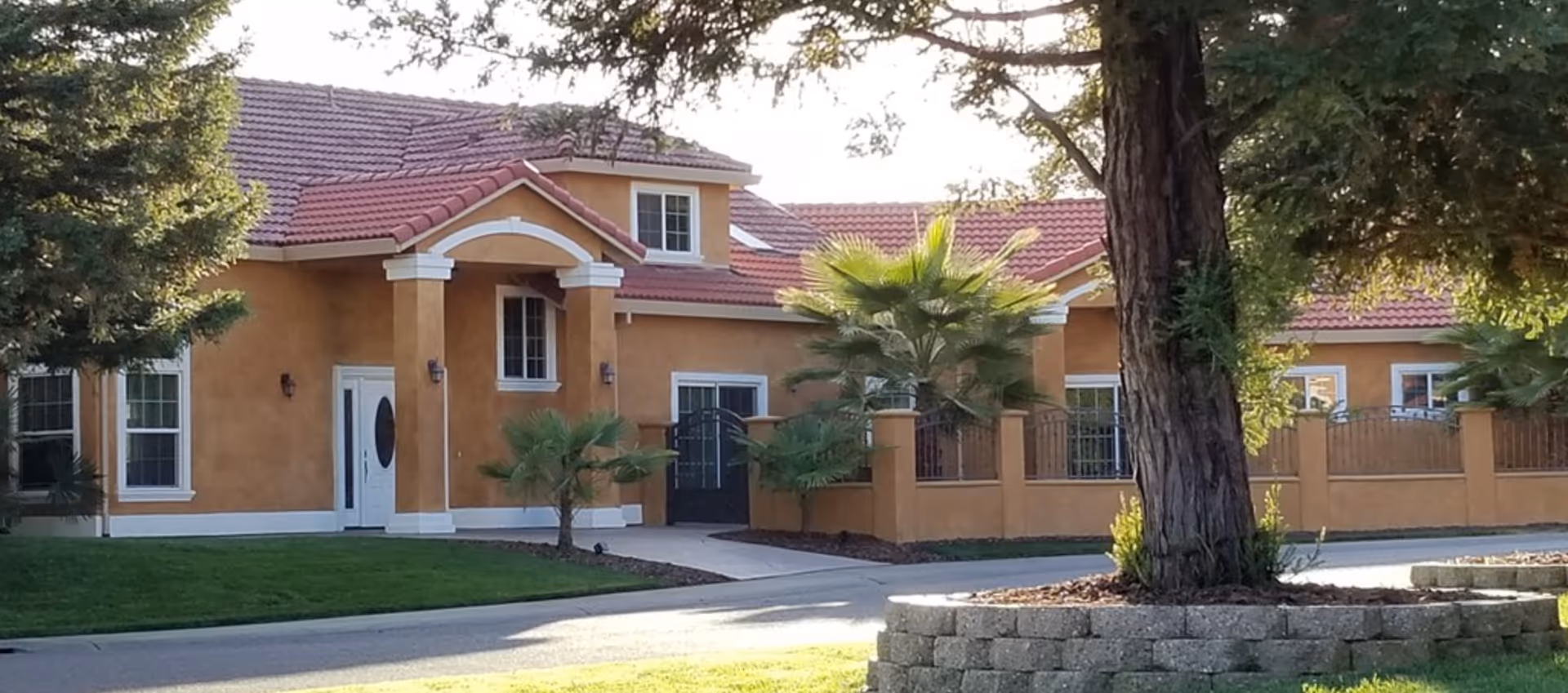 A stucco single-story building with a red tile roof, columned entrance, palm trees and a circular raised tree bed in the front yard.