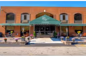 Front exterior view of Green Meadows Nursing & Rehabilitation Center showing a brick building with a green awning over the entrance. There are potted plants and flowers arranged along the walkway leading to the glass double doors.