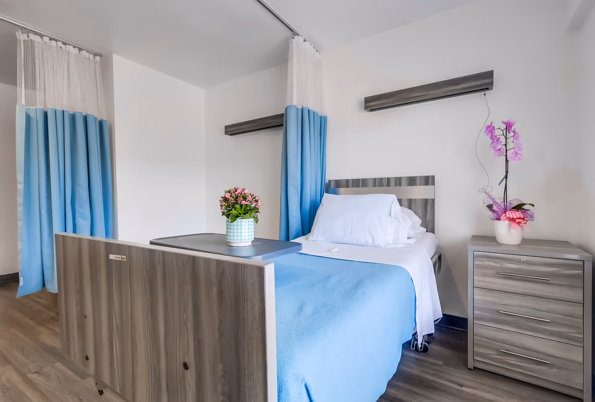 A clean and bright patient room with a single hospital bed covered in white sheets and a blue blanket. There is a bedside table with a pink orchid plant in a white pot, and a small table on the bed with a potted pink flower. Blue and white privacy curtains hang from the ceiling. The floor is wood, and the walls are white.