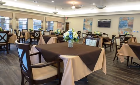 A spacious dining room with multiple tables covered in beige and brown tablecloths, each adorned with a floral centerpiece. The room has wooden chairs, large windows with blinds, and two colorful abstract paintings on the wall. A flat-screen TV is mounted on the wall between the paintings.