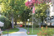 A landscaped outdoor area at Mission Care & Rehabilitation Center featuring a curved concrete walkway, green grass, trees, flowering bushes, and two American flags on flagpoles near a multi-story building.