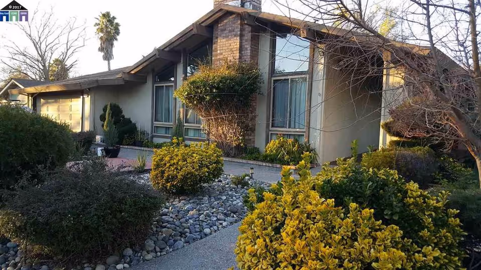 Exterior view of a single-story residential building with large windows and a brick chimney. The front yard features various bushes, shrubs, and a rock garden pathway leading to the entrance. Trees without leaves are visible, indicating a possible winter season.