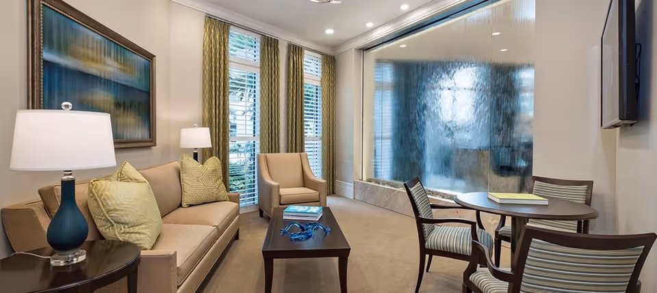 A cozy living room area with a beige sofa, two beige armchairs, a wooden coffee table with decorative items, a round table with four striped chairs, two table lamps, large windows with curtains, and a large indoor water feature on one wall.