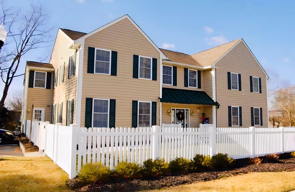 Two-story beige residential building with green shutters and a white picket fence under a clear blue sky.