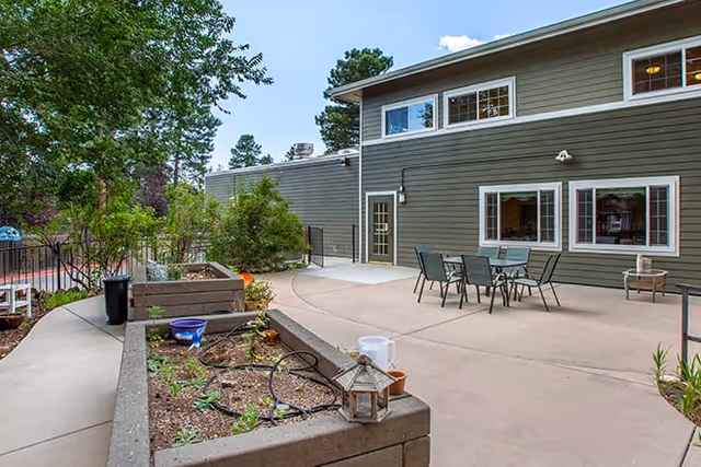 Outdoor patio area at Brookdale Flagstaff with raised garden beds, patio table and chairs, and a green building with multiple windows in the background under a partly cloudy sky.