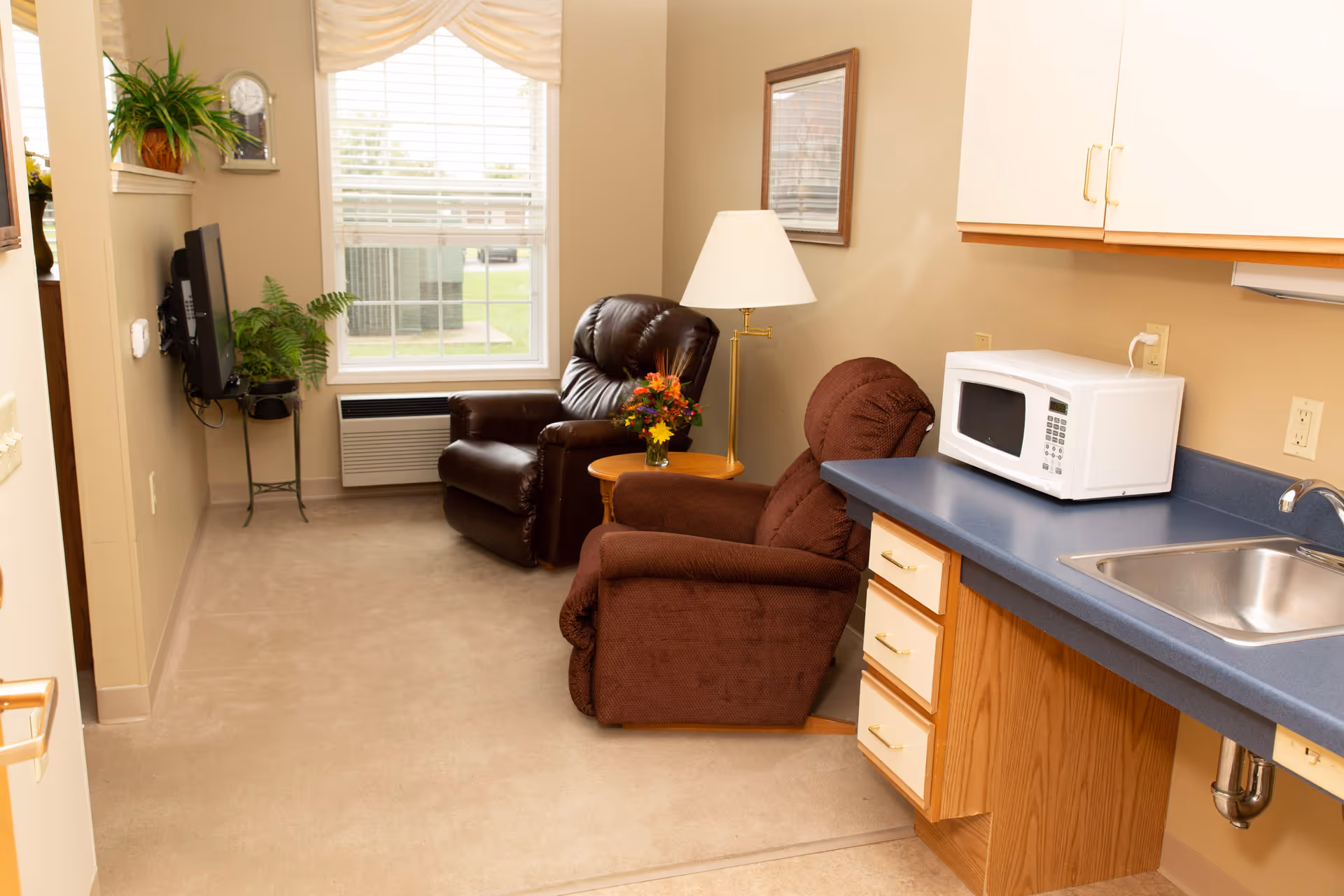 A cozy interior room with two armchairs, a small round table with a flower vase, a floor lamp, a wall-mounted TV, a window with blinds, and a kitchenette area with a microwave, sink, and cabinets.