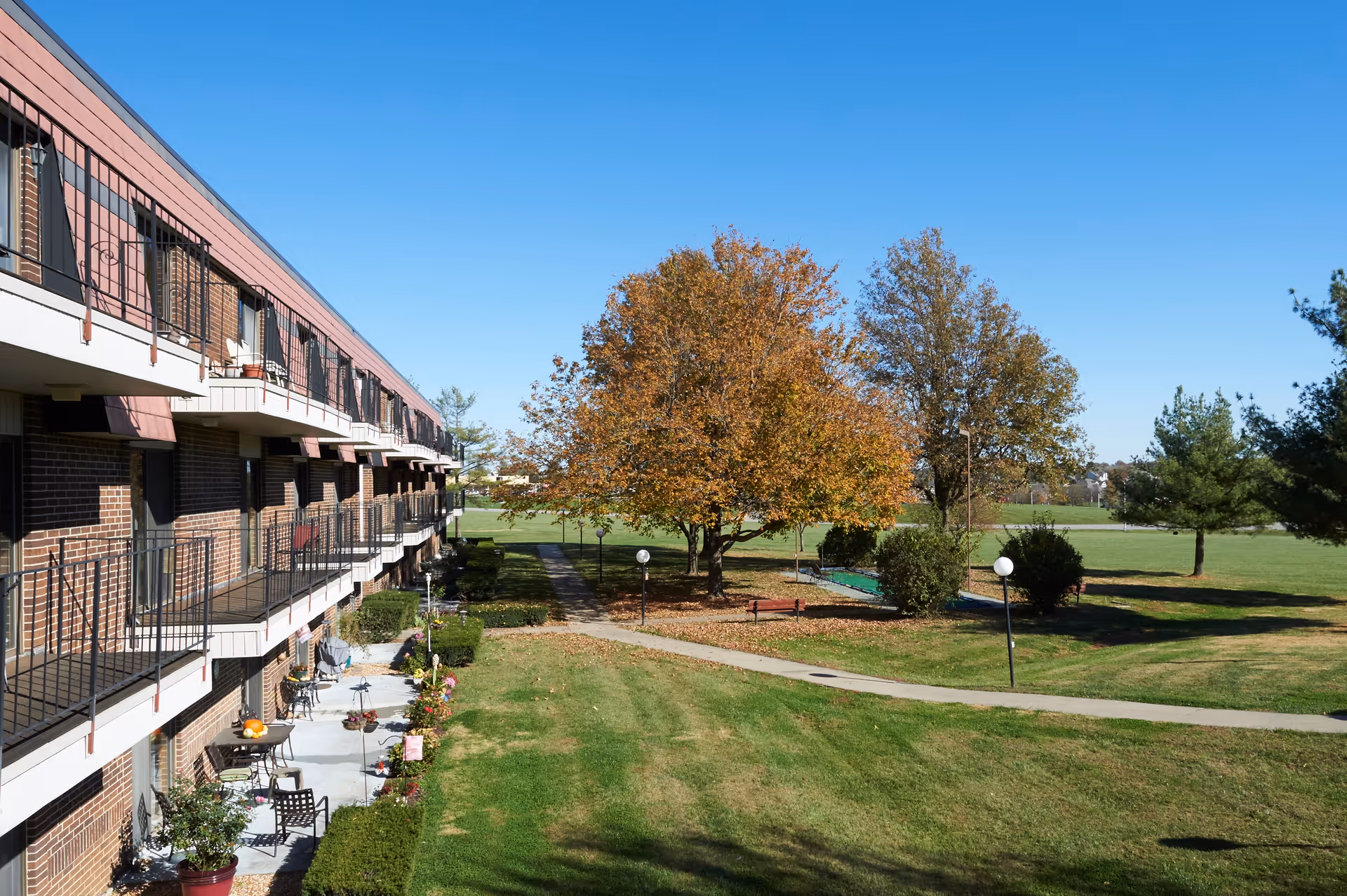 Multi-story brick senior living building with balconies overlooking a grassy courtyard, trees, and clear blue sky.