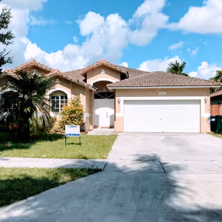Single-story house with a tiled roof, a two-car garage with a white door, and a front yard with green grass and palm trees under a blue sky with scattered clouds.