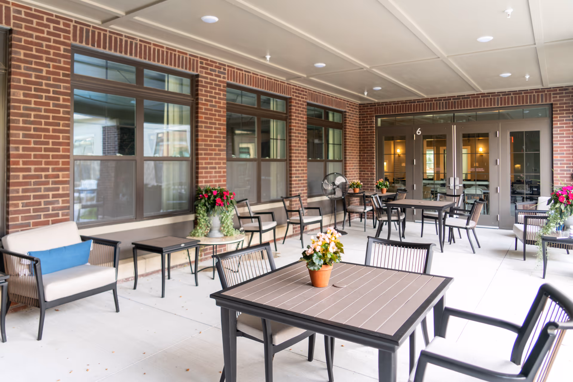 Covered outdoor patio area with tables, chairs, potted flowers and a brick building facade.