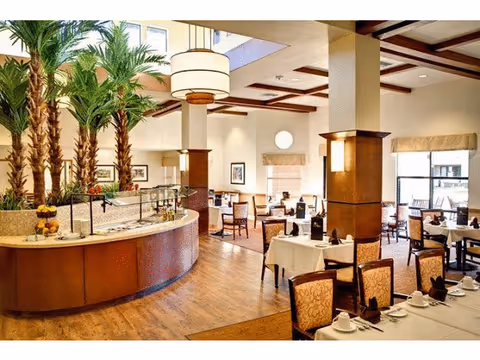A spacious dining room with multiple tables covered in white tablecloths, set with cups and napkins. The room features wooden floors, large windows allowing natural light, tall indoor palm trees, and modern light fixtures hanging from the ceiling.