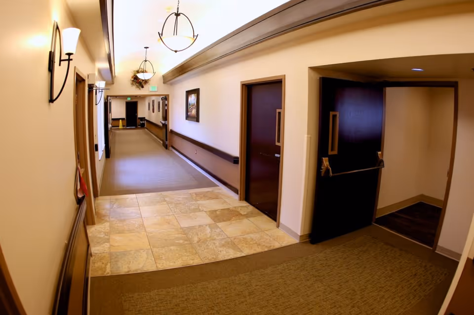 Interior hallway of a senior living facility with beige walls, tiled and carpeted floors, dark wooden doors, wall-mounted lights, ceiling light fixtures, and a framed picture on the wall.