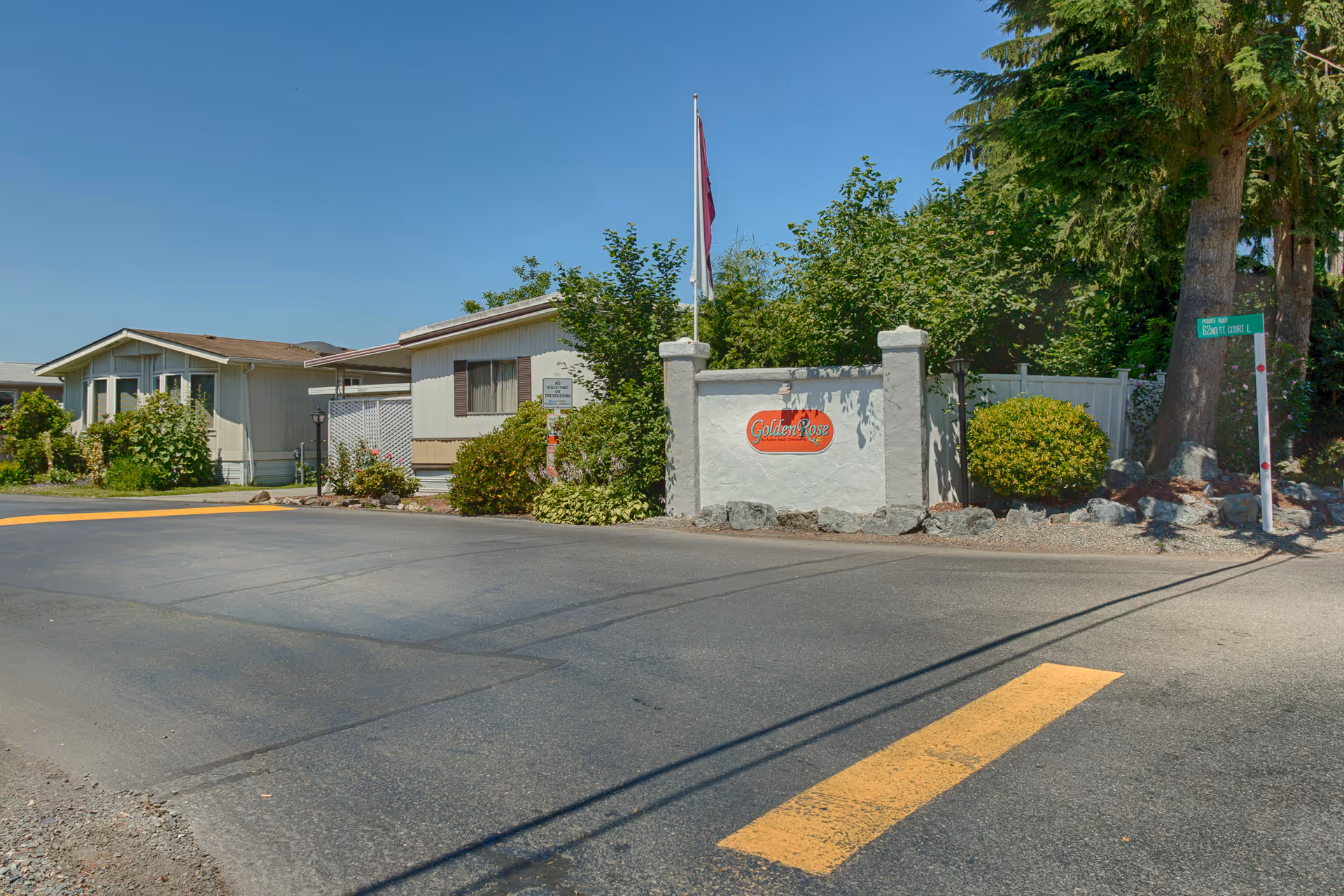 Entrance to Golden Rose 55+ Community near Tacoma Washington, showing a paved road, a white stone sign with the community's name in red, surrounded by greenery and trees, with residential buildings in the background under a clear blue sky.
