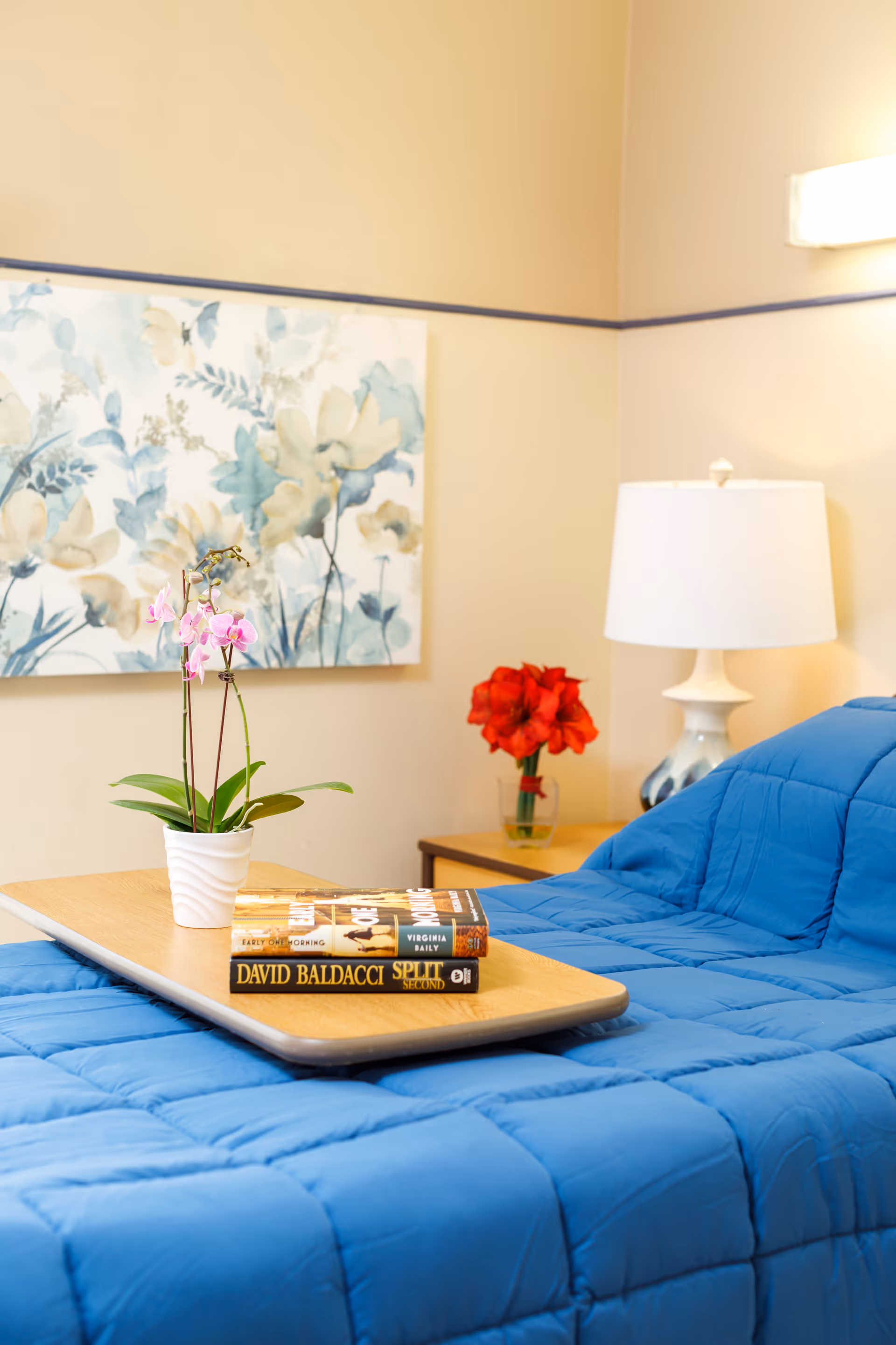 A cozy bedroom scene with a blue quilted bedspread, a wooden tray holding a small potted orchid and two books. In the background, there is a bedside table with a white lamp and a vase of red flowers, and a floral painting on the wall.