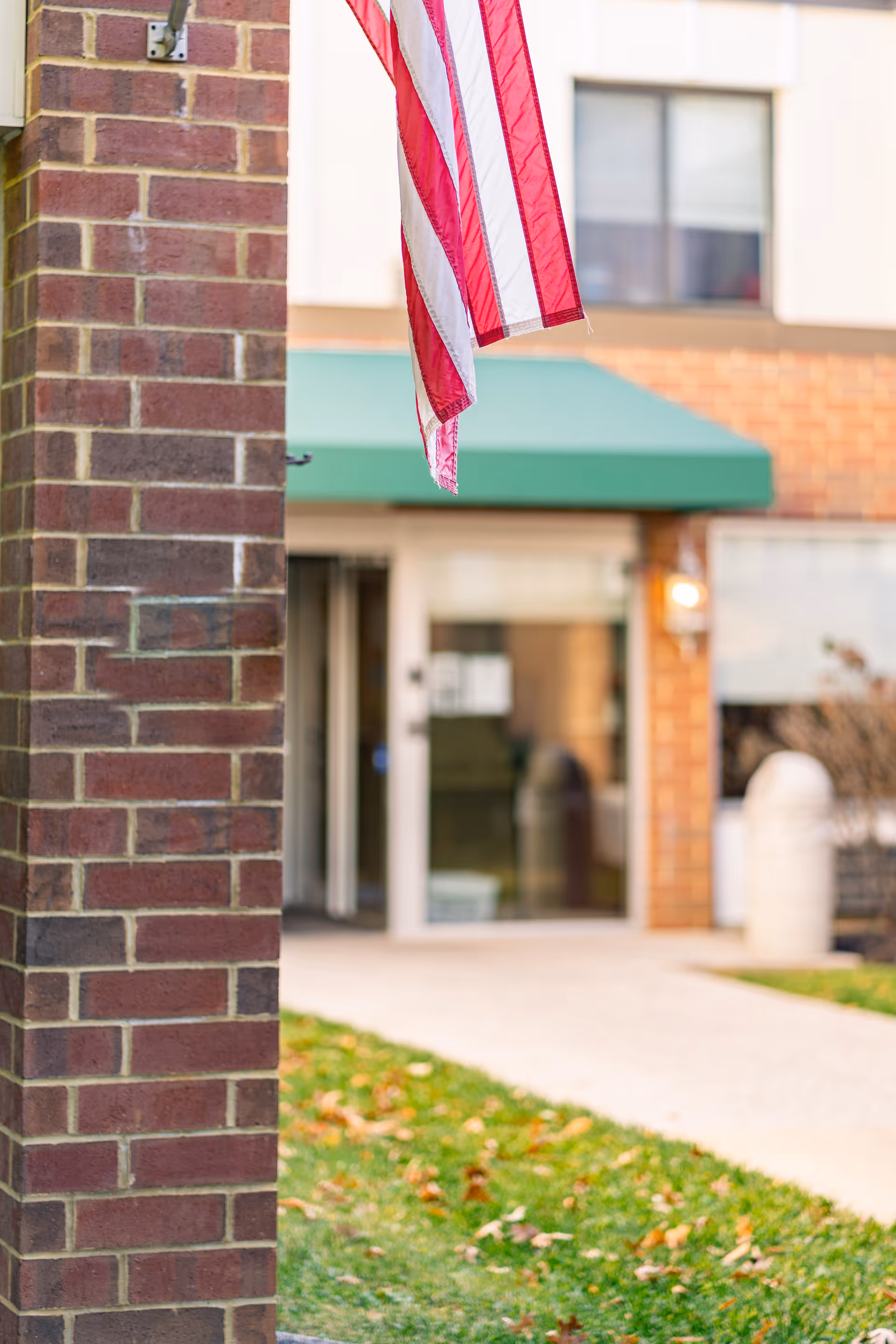Close-up of a brick pillar with an American flag hanging from it, in front of the entrance to a building with a green awning and glass doors. There is a grassy area with fallen leaves in the foreground.