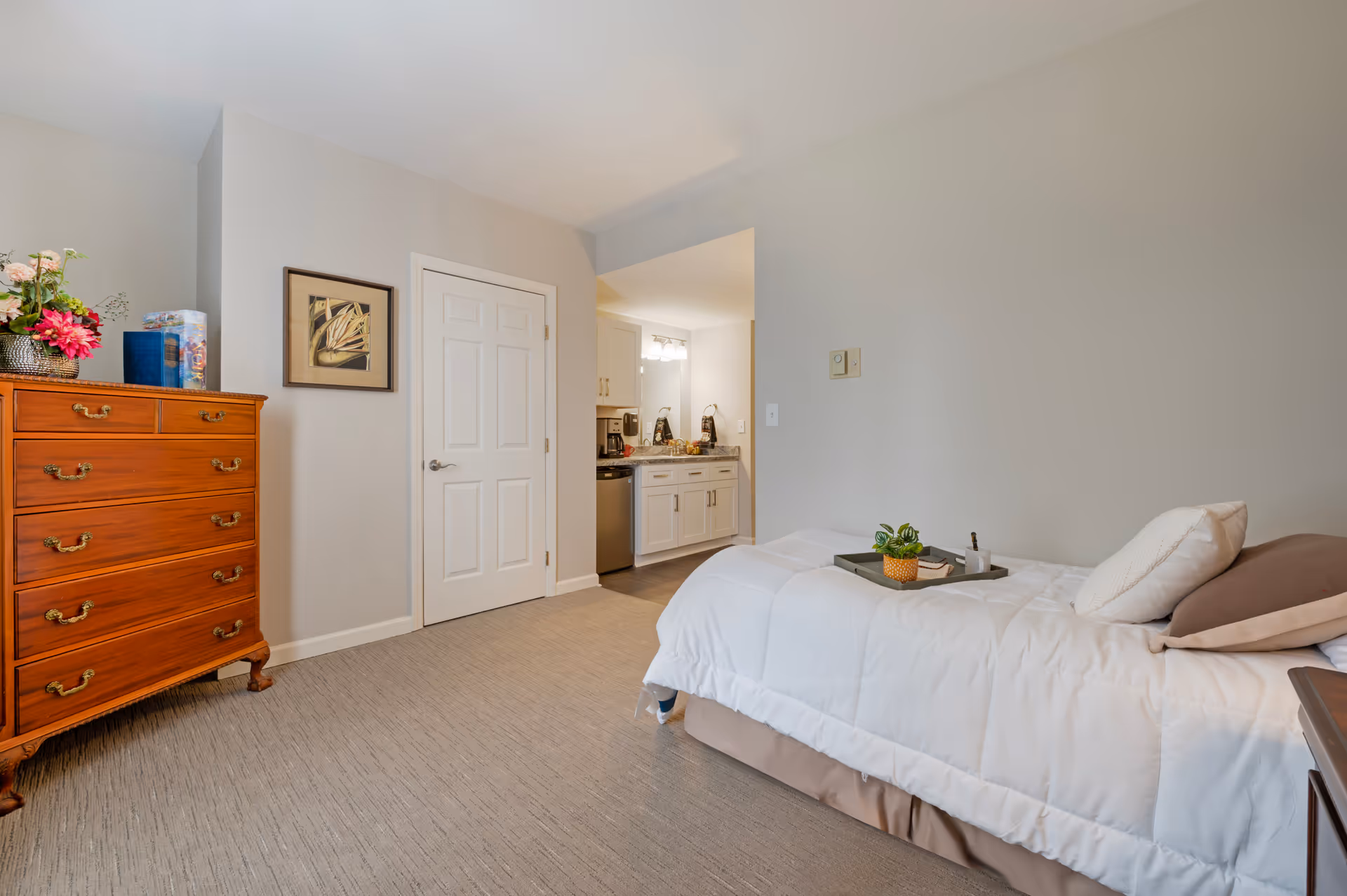 A bright furnished bedroom with a single bed made with white bedding, a wooden chest of drawers, and an open doorway leading to a small kitchenette.