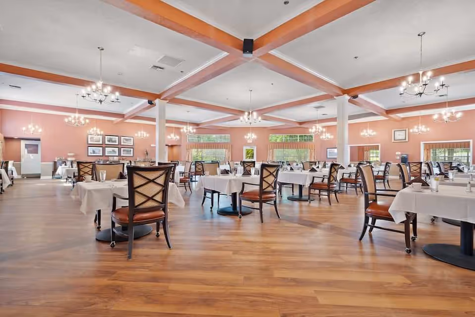 Spacious dining room with multiple tables covered in white tablecloths and set with glasses and cups. The room features wooden flooring, peach-colored walls, large windows with valances, and several chandeliers hanging from a ceiling with exposed beams.