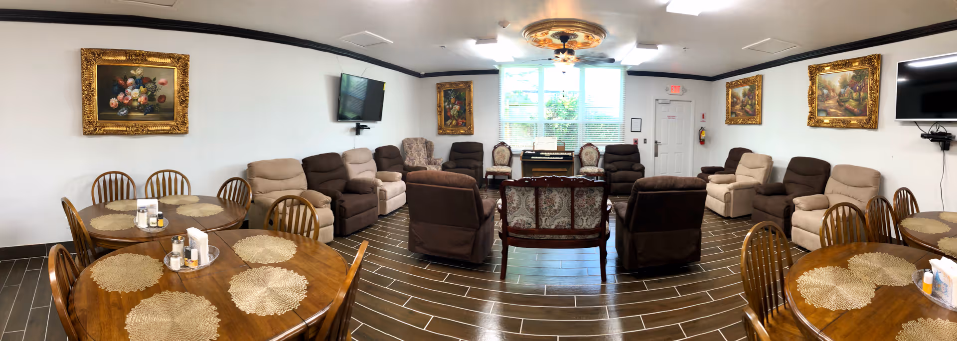 A spacious common area in an assisted living facility featuring multiple comfortable armchairs and sofas arranged around a central area. There are two round wooden tables with chairs and placemats, decorative framed paintings on the walls, two wall-mounted flat-screen TVs, and a large window letting in natural light.
