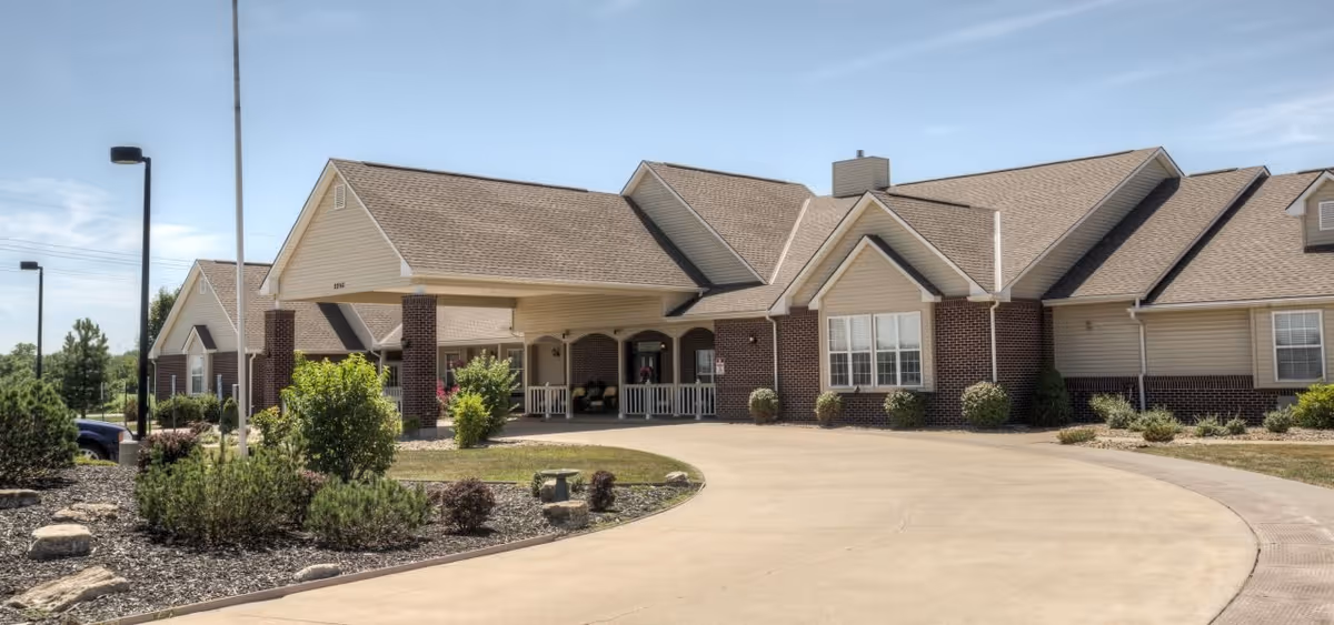 Exterior view of a single-story senior living facility building with a covered entrance, brick and beige siding, surrounded by landscaped bushes and a curved driveway under a clear sky.