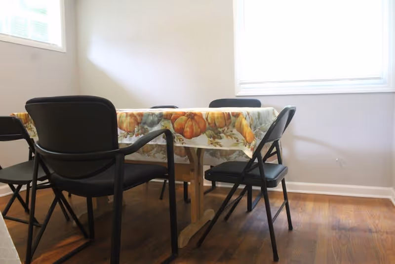 A dining area with a rectangular wooden table covered with a tablecloth featuring a pumpkin and autumn leaf pattern. Four black chairs, including folding chairs and an armchair, surround the table. The room has light-colored walls and two windows letting in natural light.