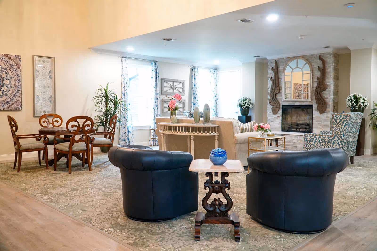 A cozy living room area with two black leather armchairs facing a small wooden table with a blue decorative item. Behind them is a beige sofa with a white console table and decorative vases. To the right, there is a patterned armchair and a stone fireplace with a mirror and wooden wall decorations. On the left side, there is a wooden dining table with four chairs and framed artwork on the wall. The room is well-lit with natural light coming through windows with floral curtains.