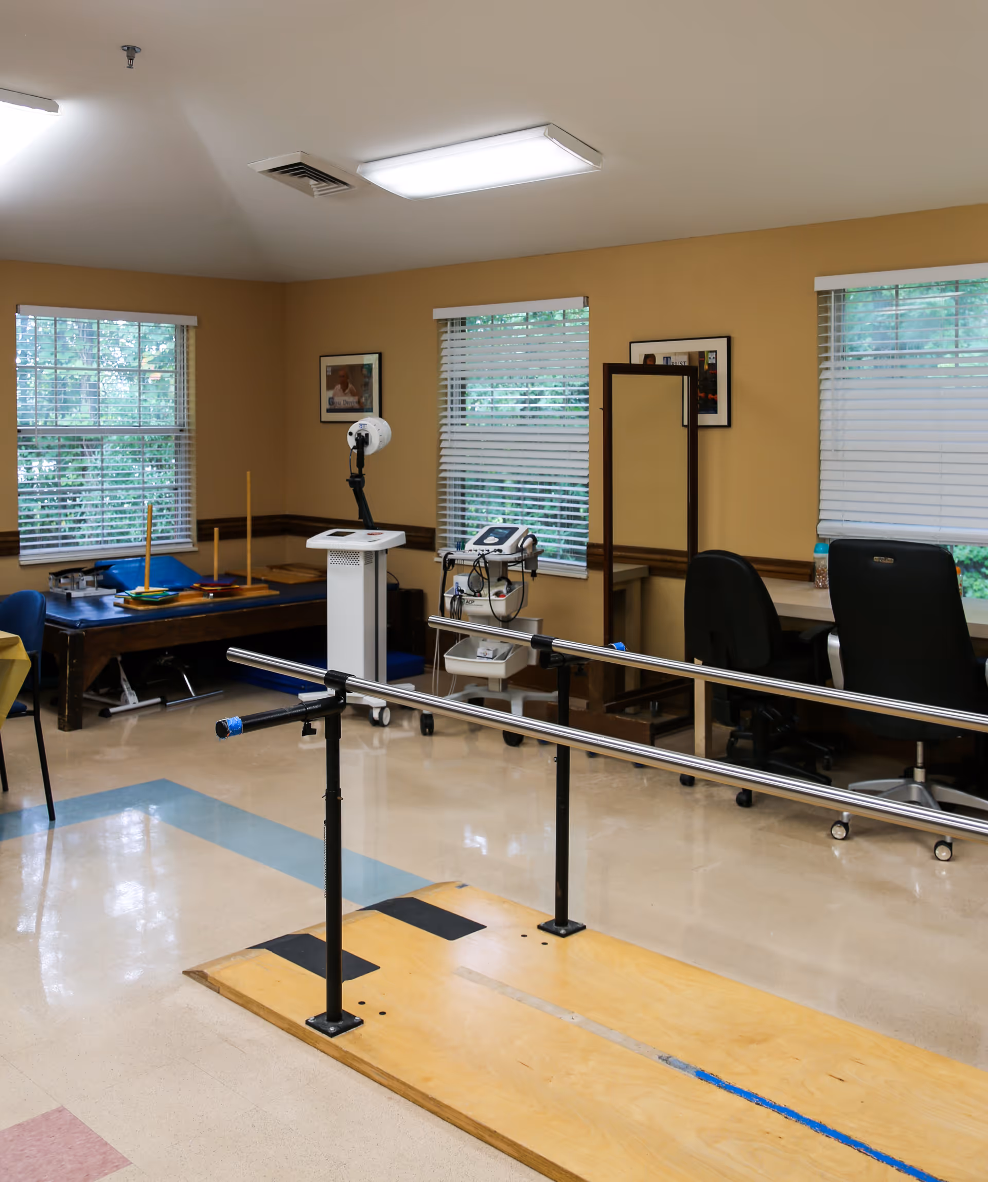 A rehabilitation therapy room with parallel walking bars, medical equipment, desks and chairs beneath windows.