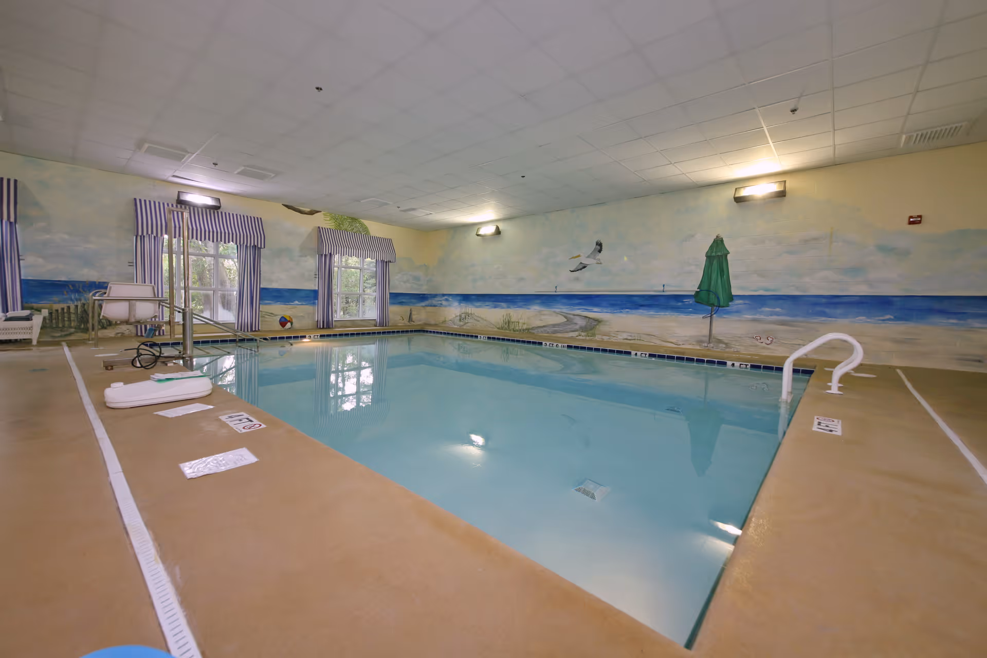 Indoor swimming pool with a beach-themed wall mural, pool lift, and striped window valances.