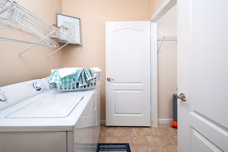 Laundry room with a white washing machine and dryer side by side, a laundry basket with folded clothes on top, beige walls, a white door slightly open, and wire shelving above the machines.
