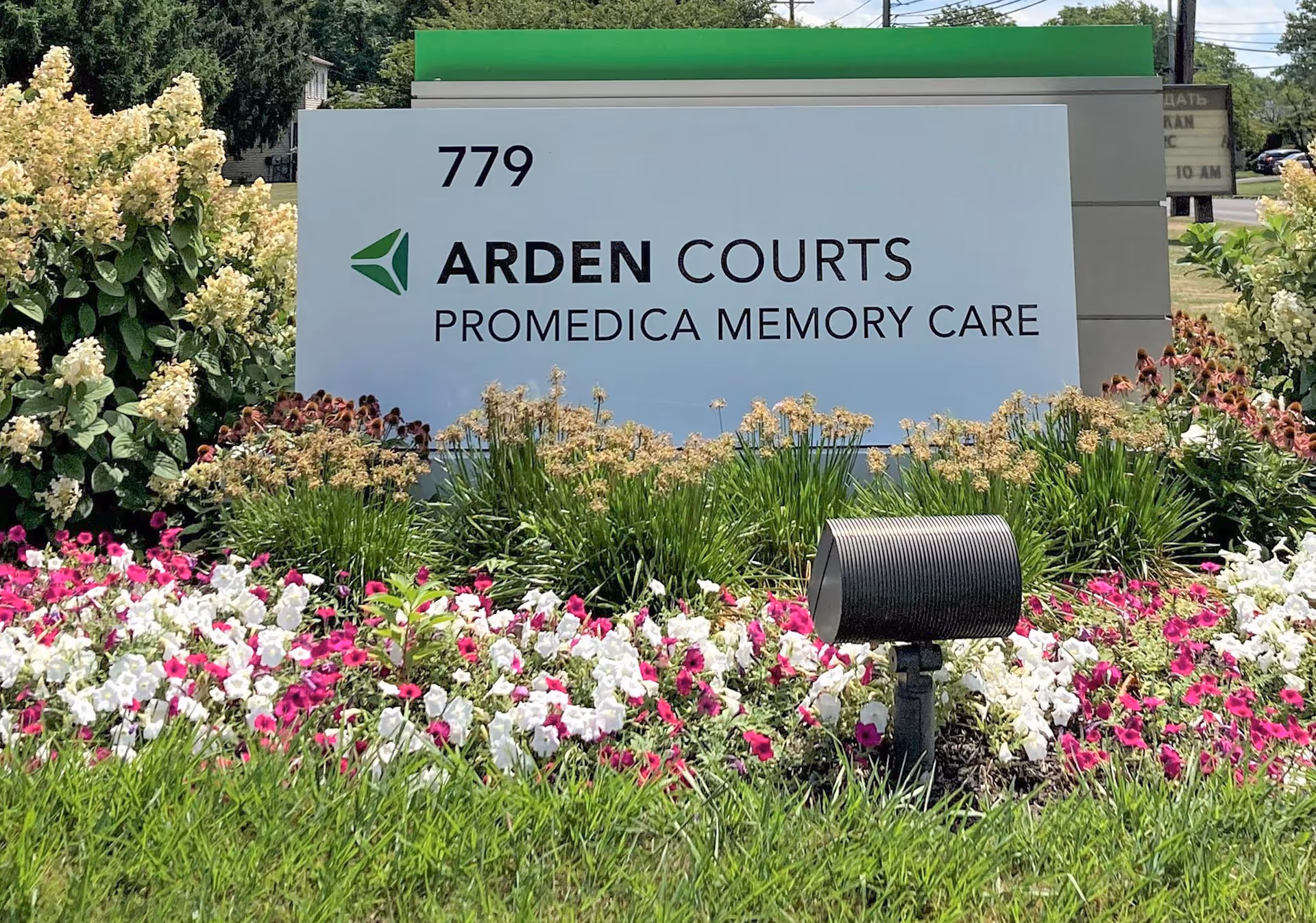 Outdoor sign for Arden Courts ProMedica Memory Care Community surrounded by colorful flowers and greenery.