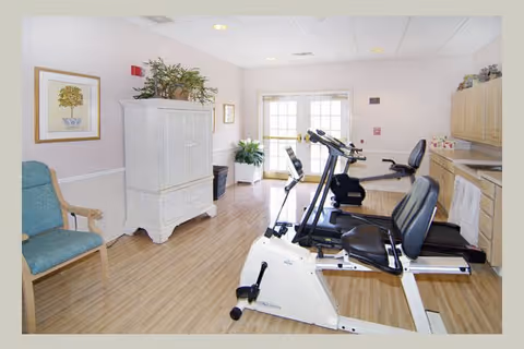 A bright exercise room in a senior living facility featuring two recumbent exercise bikes on a wooden floor. The room has light-colored walls, a white cabinet with plants on top, framed artwork, a blue upholstered chair, and large windows letting in natural light.