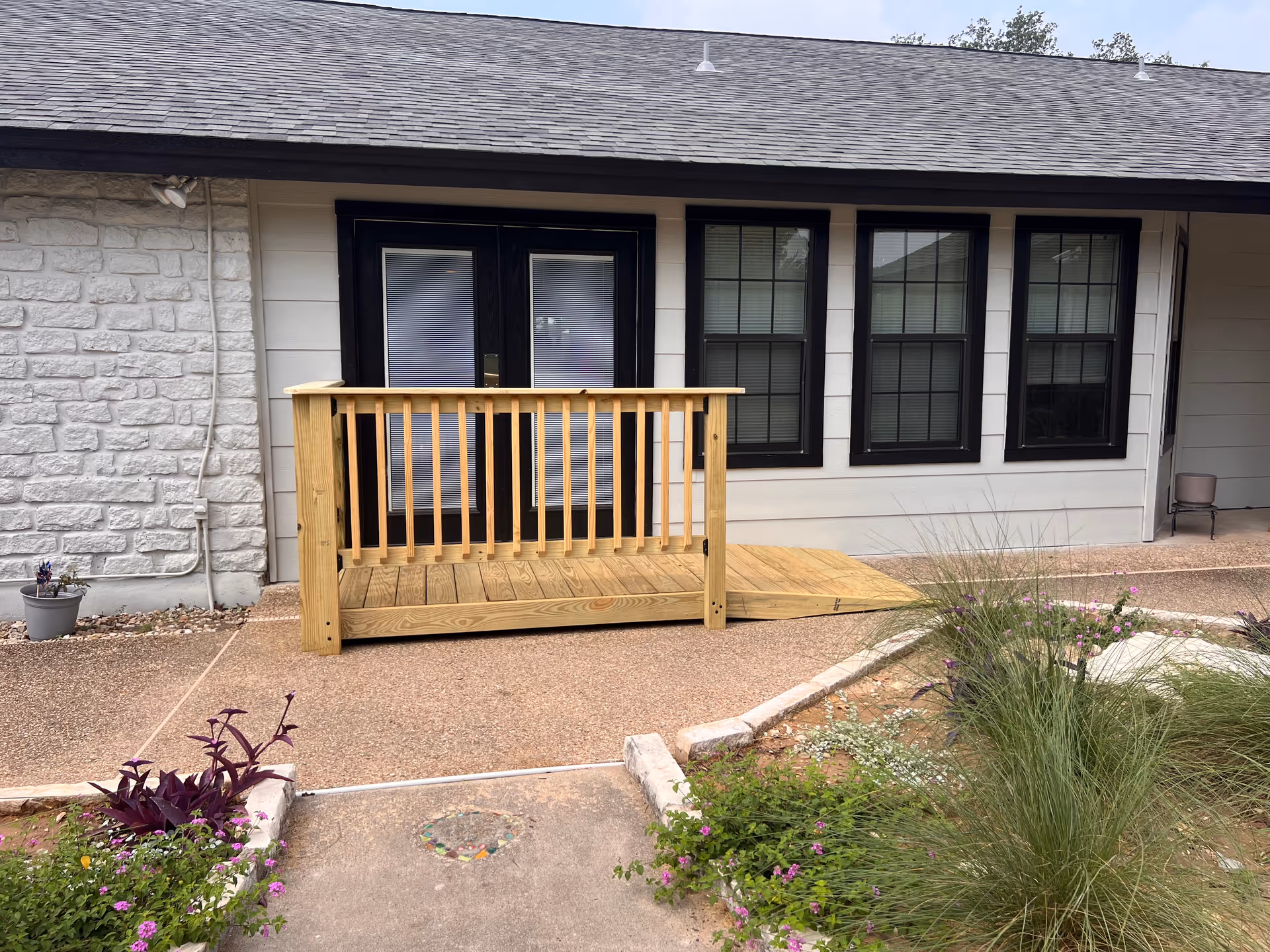 Small wooden ramp and railing leading to double doors and several windows on the front exterior of a senior living building with landscaping in the foreground.