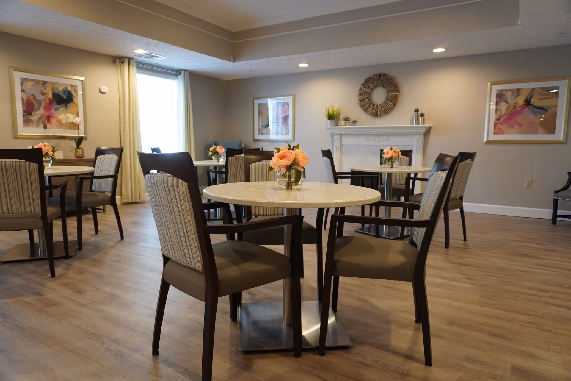 A dining room with round tables and cushioned chairs arranged neatly. Each table has a small vase with pink flowers. The room features light wood flooring, neutral-colored walls, framed abstract artwork, a window with light curtains, and a decorative fireplace with a wreath and small plants on the mantel.