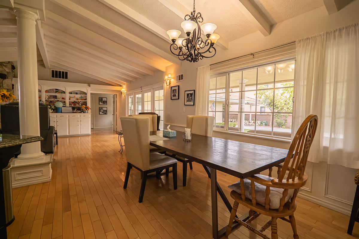 A bright dining room with a wooden table surrounded by six chairs, including a wooden armchair and upholstered chairs. The room features large windows with white curtains, a chandelier hanging from a white beamed ceiling, and hardwood floors. In the background, there is a white cabinet with decorative items and framed pictures on the walls.