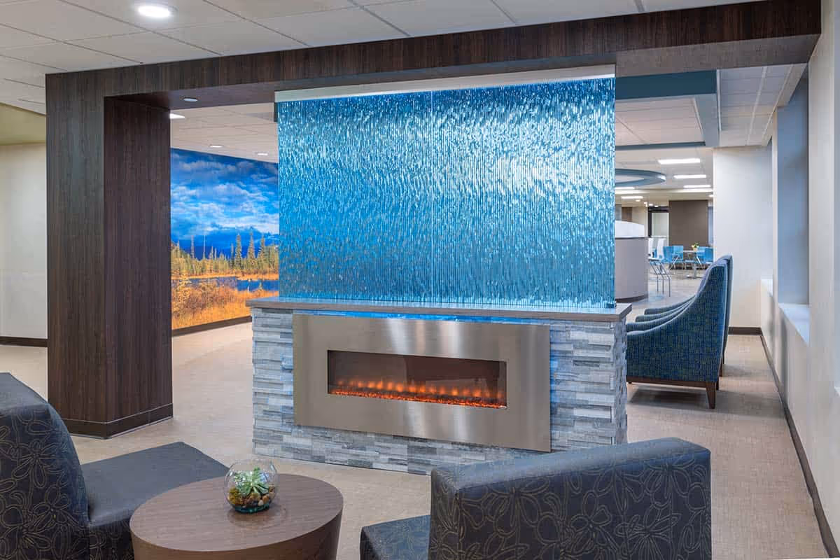 A modern senior living facility lobby with a stone fireplace, a blue glass divider, and blue upholstered seating.